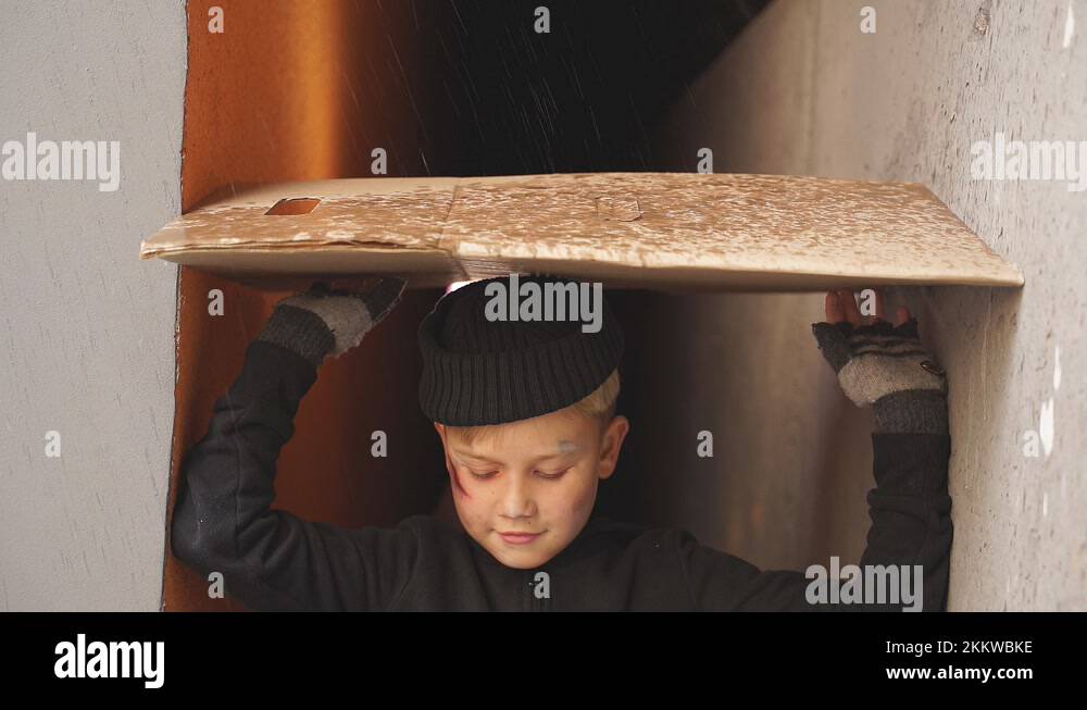 Poor orphan boy shelters from the rain under cardboard, crying Stock