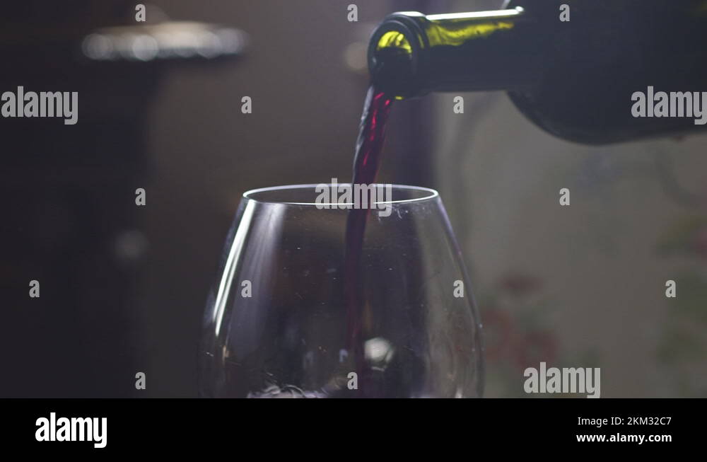 A man pours red wine into a glass from a green bottle in a dark room ...