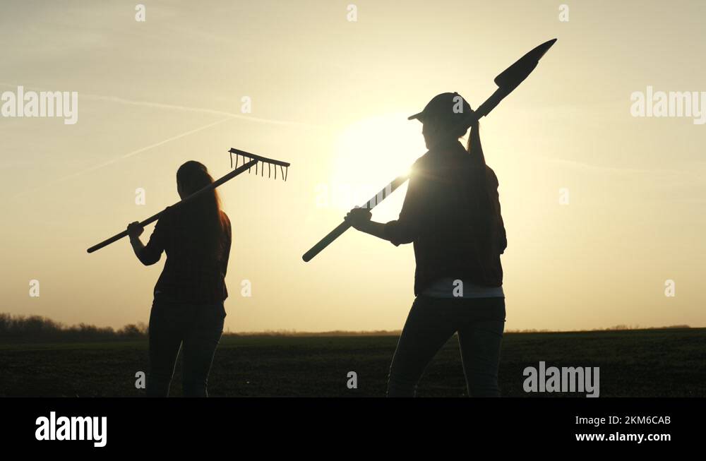 Farmer girls walk across the field with a shovel and a rake on their ...