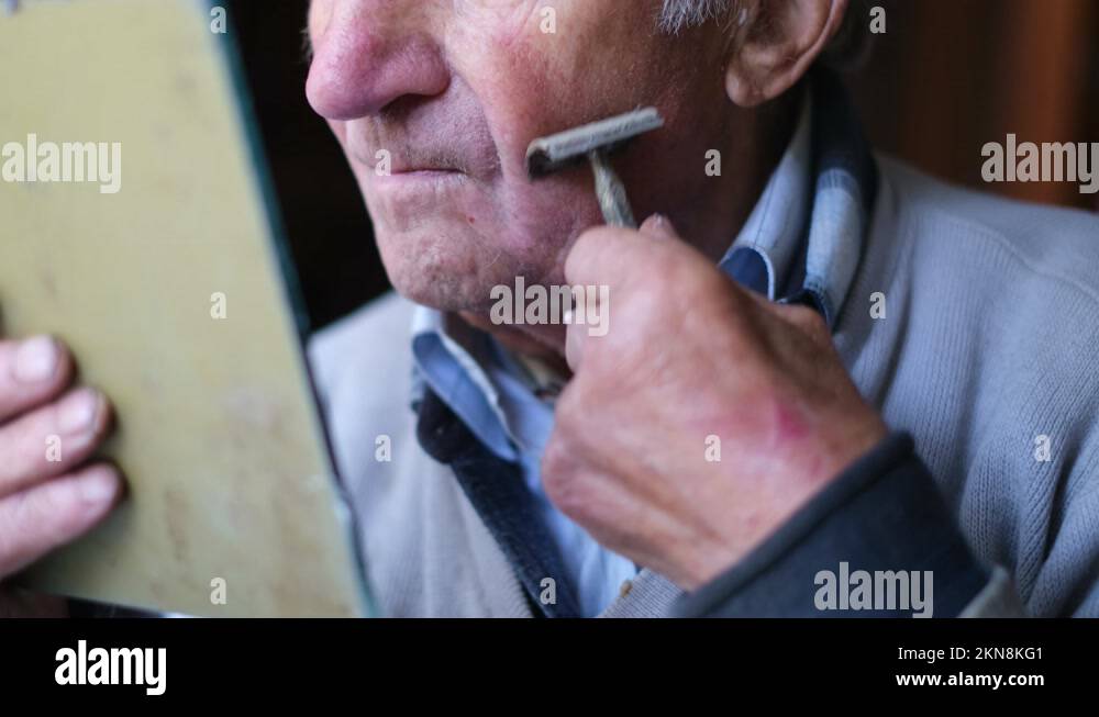 An elderly man shaves his face in front of a mirror. The pensioner