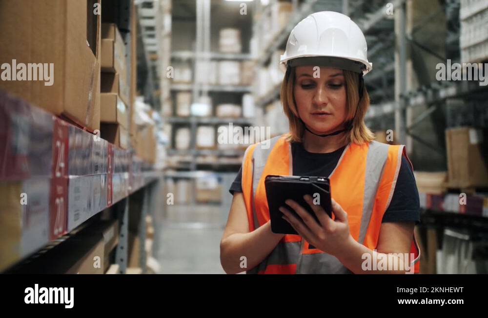 Female worker warehouse wearing hard hat and safety vest checks stock