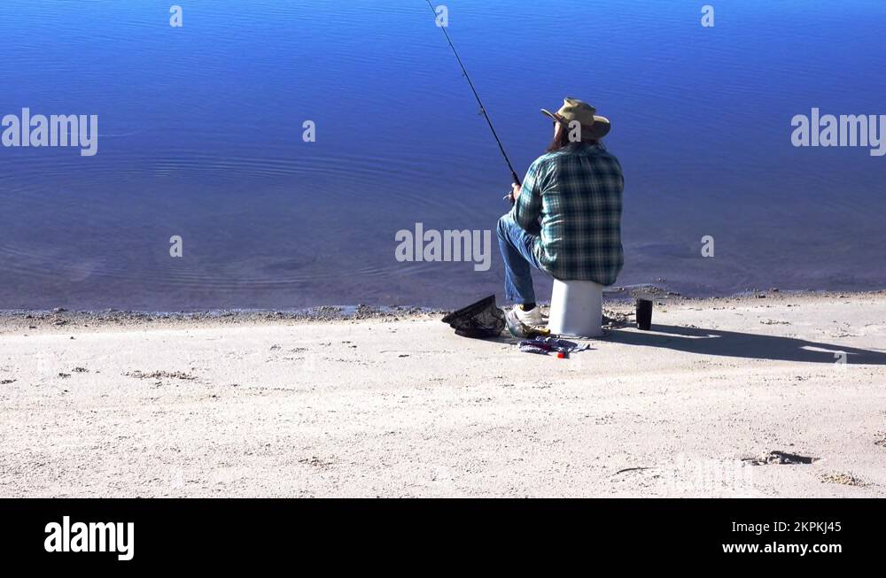 Fisherman sits on an upside down bucket on a beach and casts out his