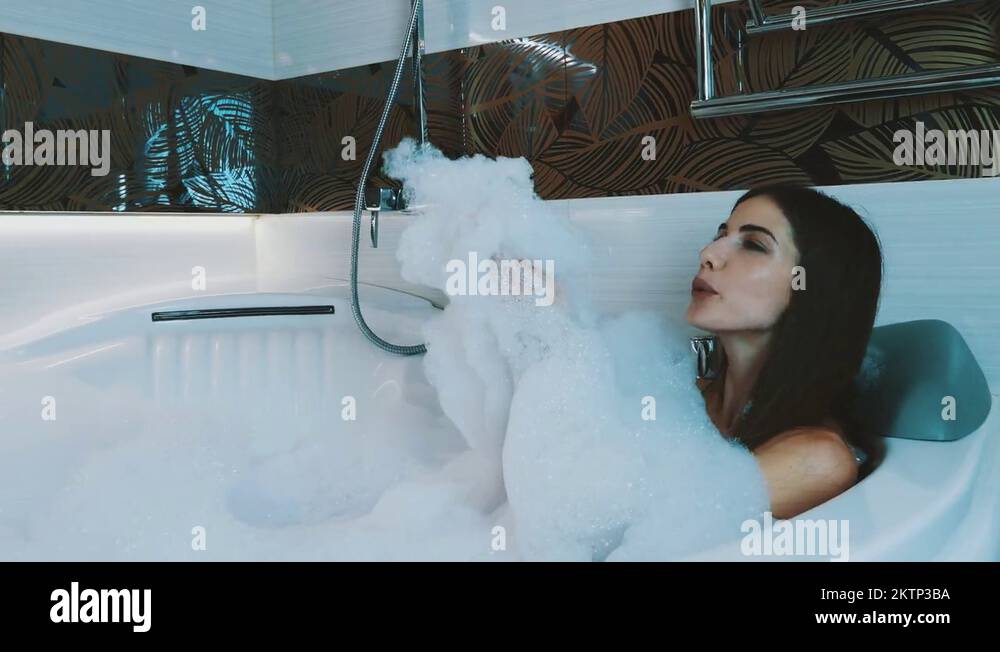 Brunette young girl taking bath full of foam in bathroom. Blow bubbles