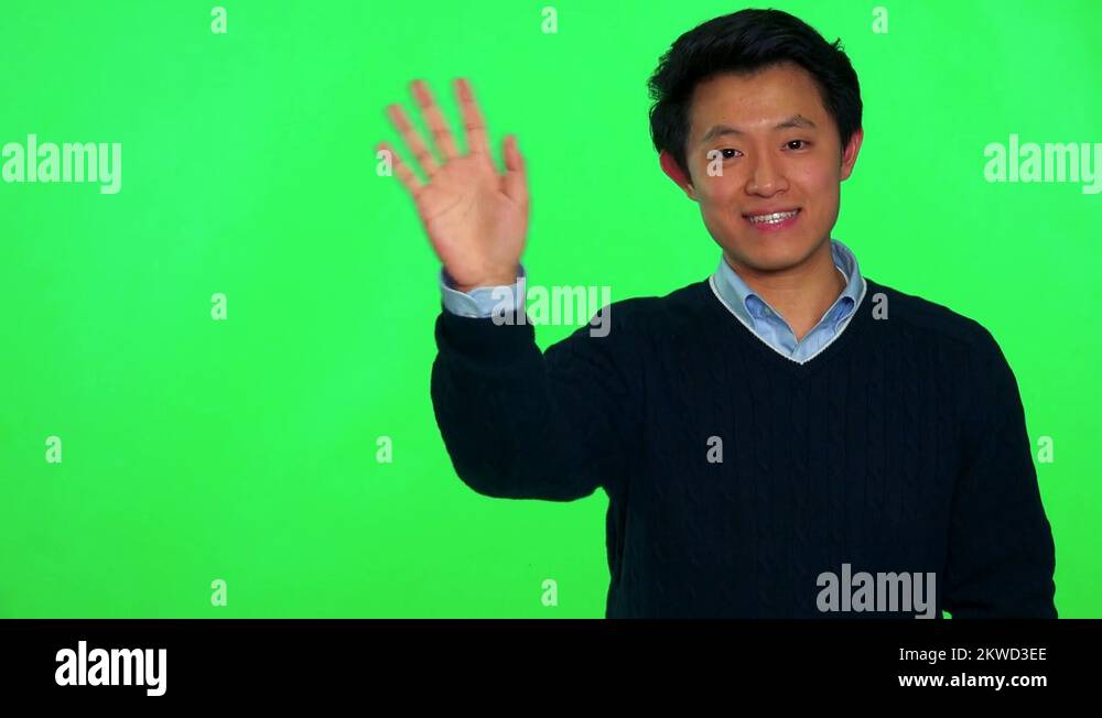A young Asian man waves and smiles at the camera - green screen studio ...
