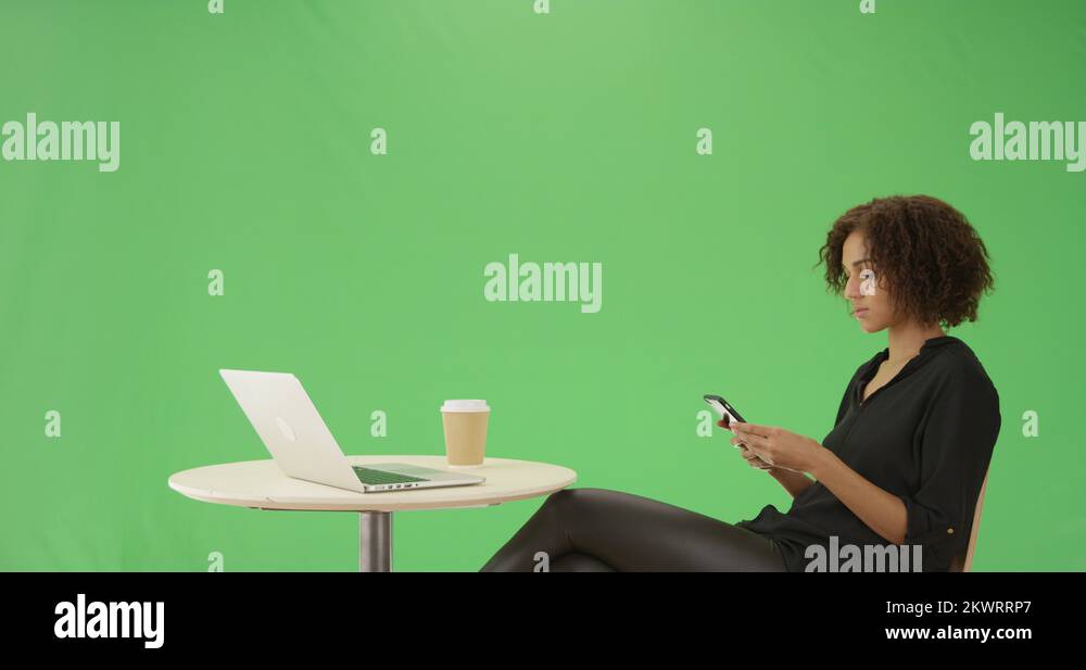 A young black woman sits at a caf table on her phone on green screen ...