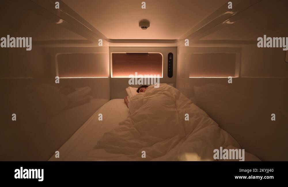 Man sleeps inside a clean modern capsule hotel room with lights and