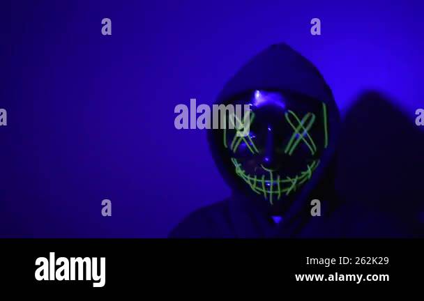 A hooded figure wearing a glowing green neon mask moves through the ...