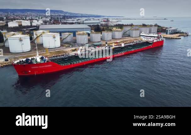 A red tanker ship docked at an industrial port with large storage tanks ...