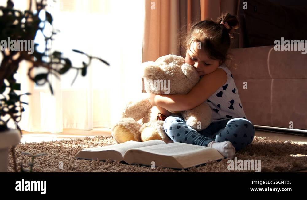 Little girl sitting on the floor reading a book hugging toy teddy bear ...