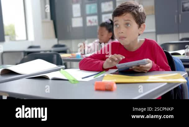 In school classroom, young biracial boy holding a tablet looks up with ...