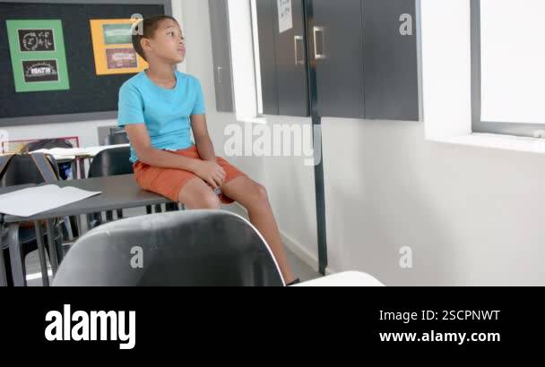 Biracial boy sits on a desk in a classroom, looking towards the door ...