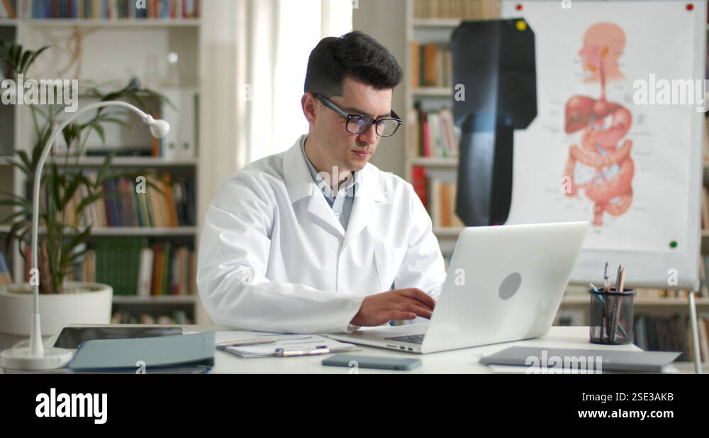 Young man in white rob filling up medical form while typing at laptop ...