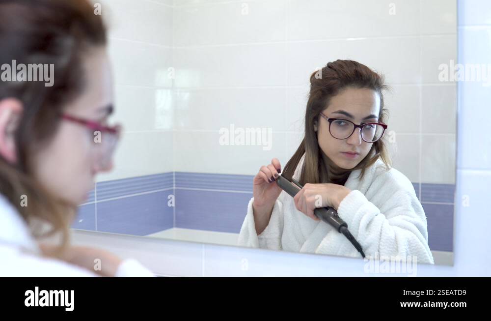 A young woman in glasses straightens her hair with a curling iron in ...
