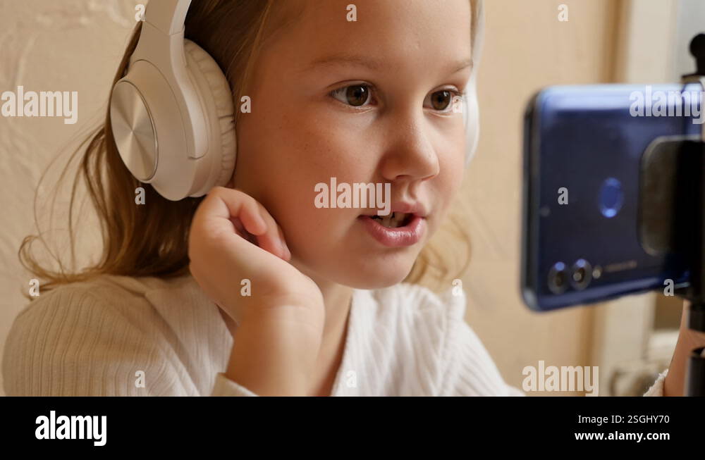a cute teenage girl in white big headphones is talking on a video call ...