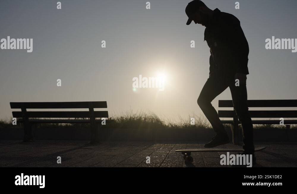 Guy chilling on bench backlit at sunset jumps on skateboard and rides ...
