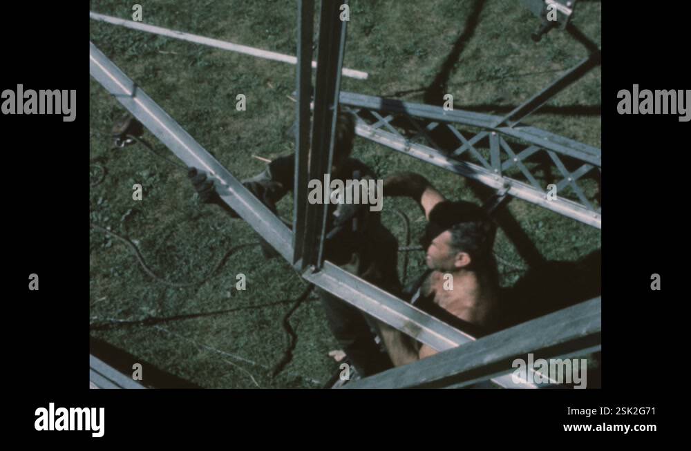 1950s: Men line up the metal framing of a Ferris wheel. A man hammers ...