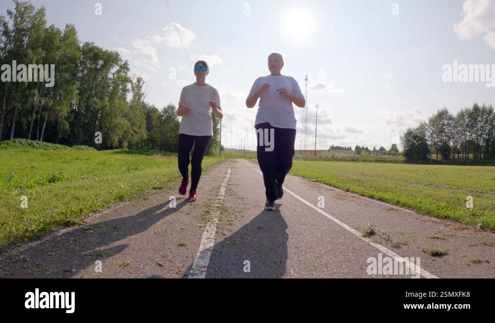 Two women are running on a paved path on a sunny day. They are wearing ...