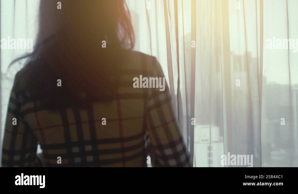 Young pretty brunette woman open cutrains stands by the window drinks ...