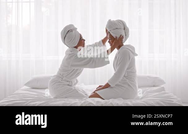 Two African American sisters sit closely on a bed, draped in cozy white ...
