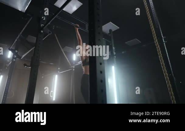 A sporty young woman pulls herself up on a horizontal bar in a dark gym ...