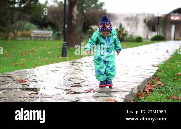 Little girl in rubber boots walks through puddles on a paved path in ...