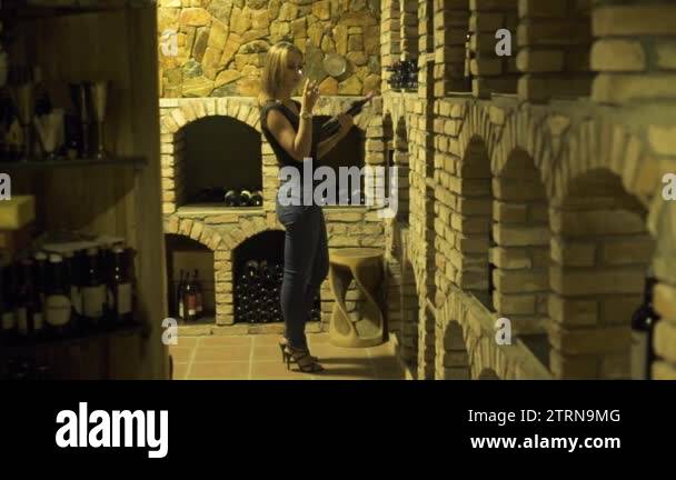 Woman drinking red wine from glass and holding bottle in hand in cellar ...