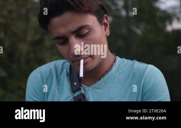 Handsome hispanic man takes a cigarette and light it up. Tired guy smokes a  cigarette wearing blue shirt. Man has beauty spot birthmark right over lip