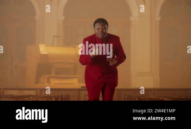 Smiling gospel performer in red suit standing at row of wooden church