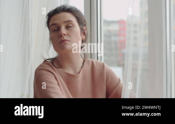 Young sad woman sitting on sill and looking through the window while ...