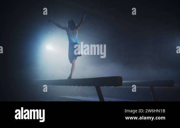 young female gymnast is training on balance beam in dark sports hall ...