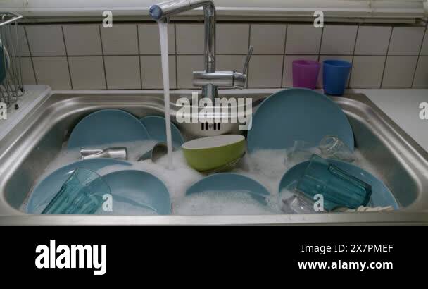 Piled dishes of various colors soak in soapy water under a running ...