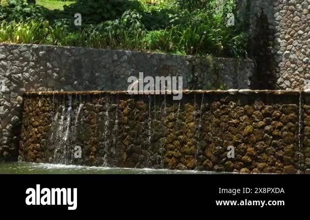 Water cascading down textured stone wall into serene pond in lush green ...