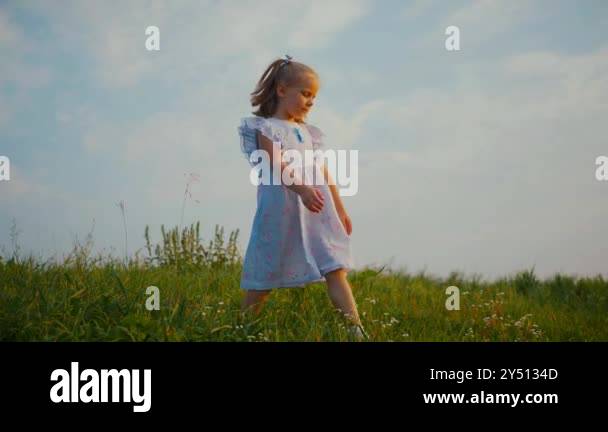 A little girl in a blue dress standing happily in a sunlit field. A ...