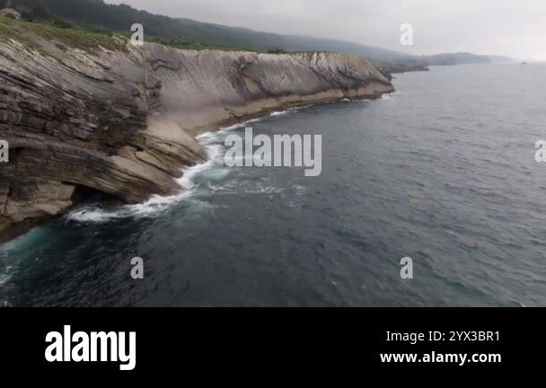 Ocean waves crash against rugged cliffs under a cloudy sky, creating a ...