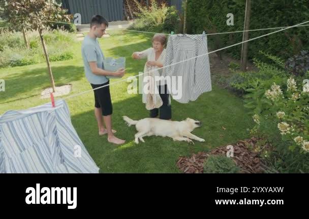 Image showcasing A young man and an older woman hang laundry on a clothesline in a green ...