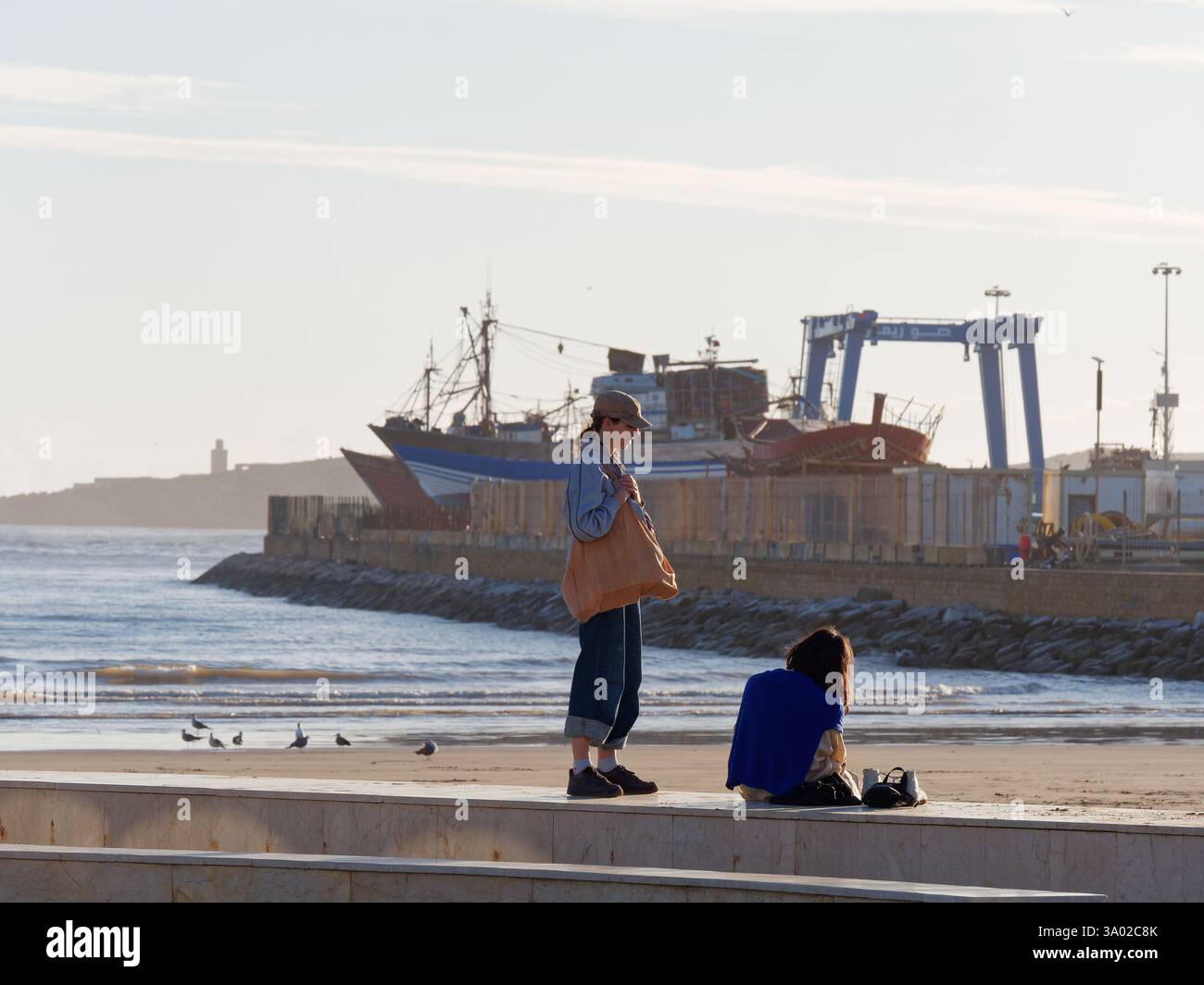 Shipyard by the sandy beach with two female friends on the promenade wall in foreground in Essaouria, Morocco. Feb/Mar 2025 Stock Photo