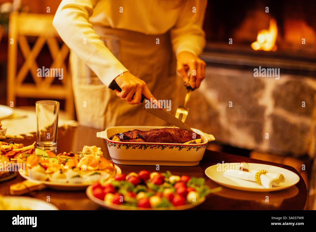 Close-up of a woman cutting a baked duck for a festive dinner. A table with delicious Christmas dishes. The lady cuts off pieces of meat. A fireplace Stock Photo