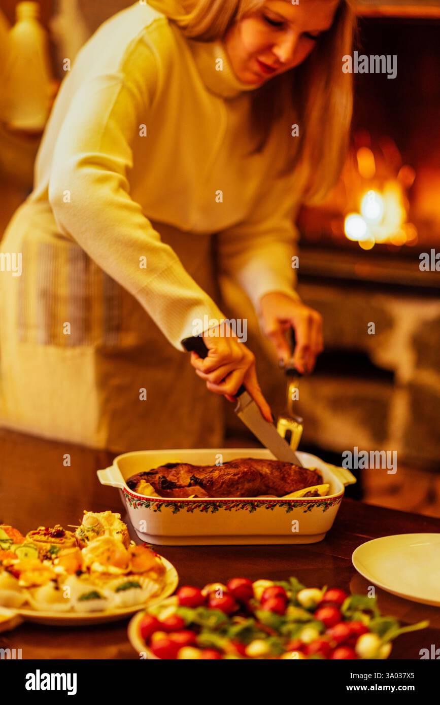 Close-up of a woman cutting a baked duck for a festive dinner. A table with delicious Christmas dishes. The lady cuts off pieces of meat. A fireplace Stock Photo