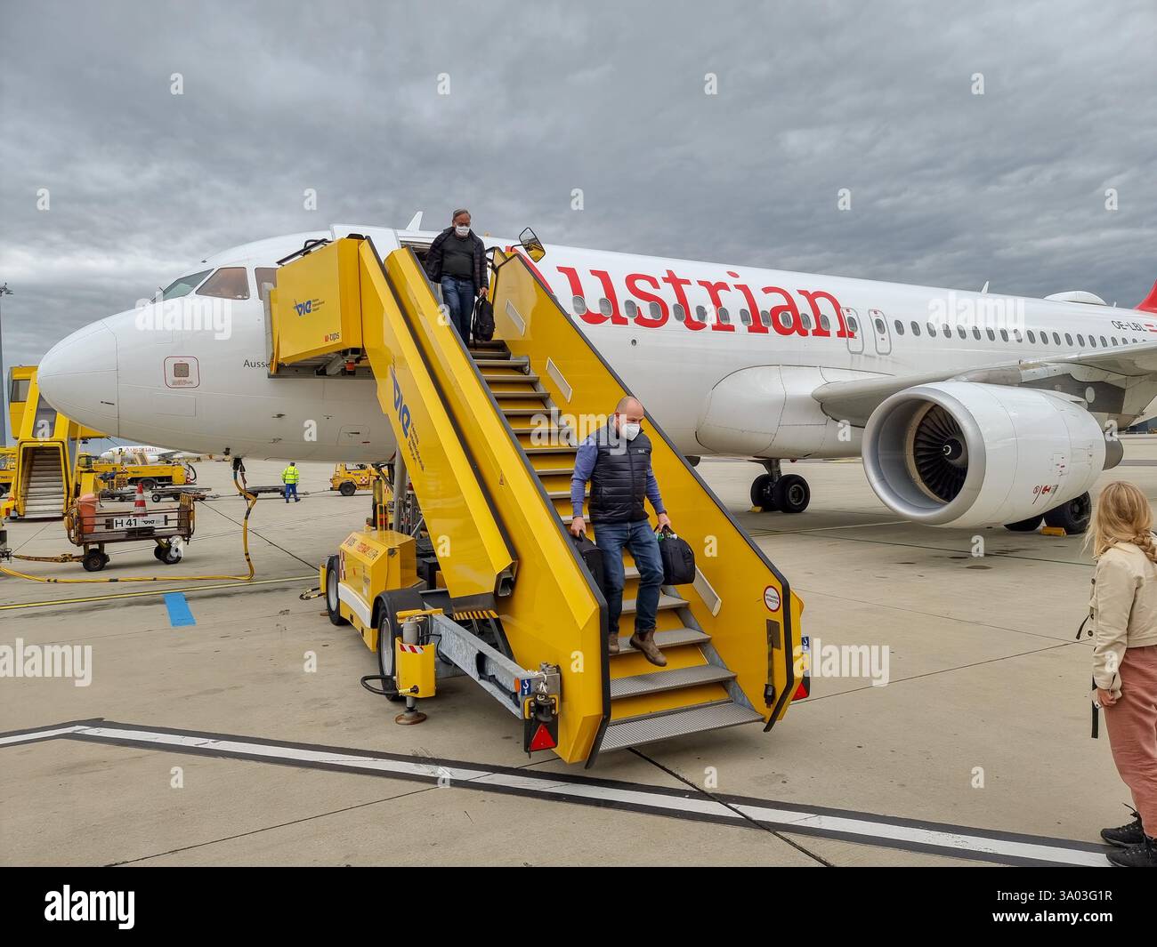 Two passengers with handbags & wearing facemasks descending from yellow stairs from an Austrian Airlines airplane onto the tarmac; leaving the plane - Smartphone Captured Stock Image