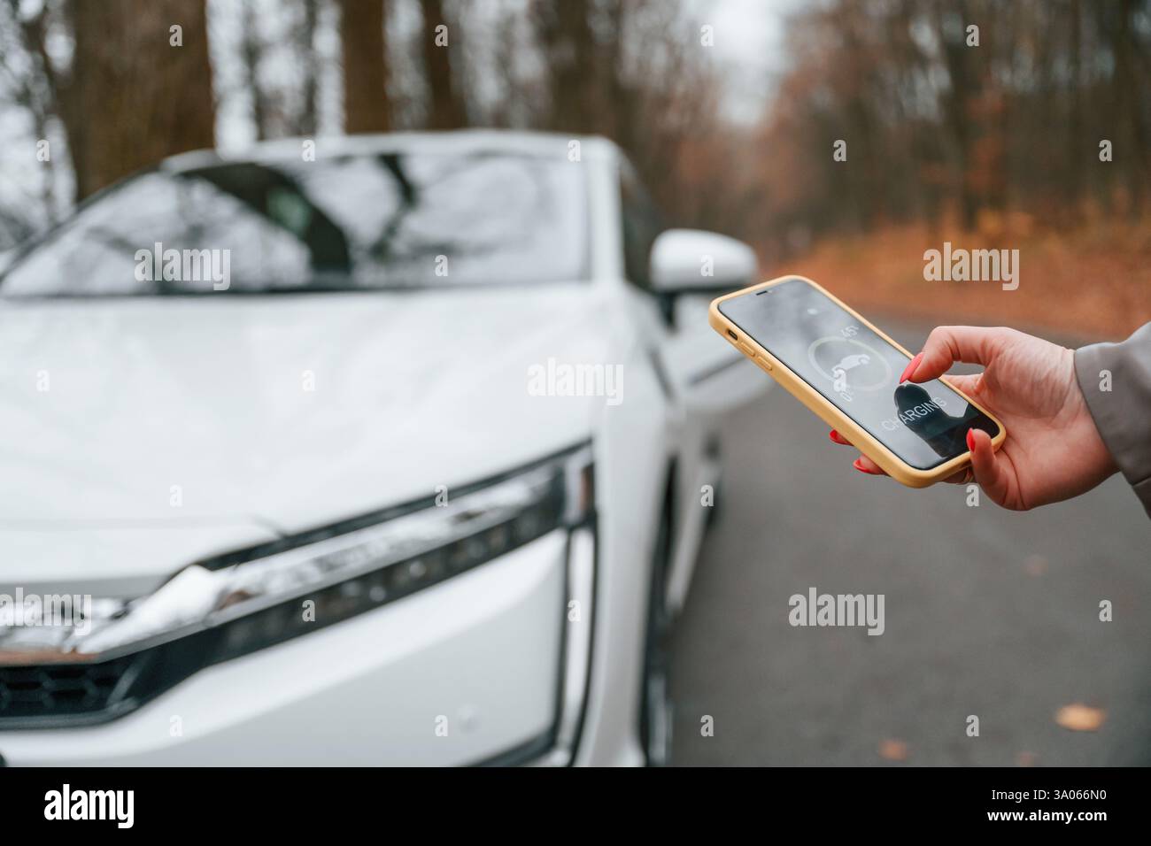 With smartphone in hand. Charging process. Woman is standing near her electric car outdoors on the road. Stock Photo