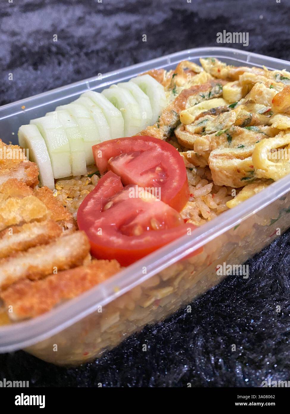 A delicious homemade bento featuring crispy chicken katsu, herb omelette, fried rice, fresh cucumber, and tomatoes. Perfect for meal prep, lunch ideas - Smartphone Captured Stock Image