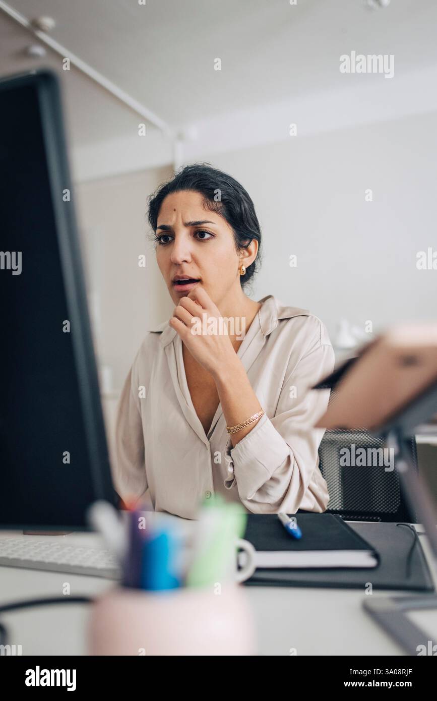Confused female creative professional using desktop PC while sitting at desk in office Stock Photo