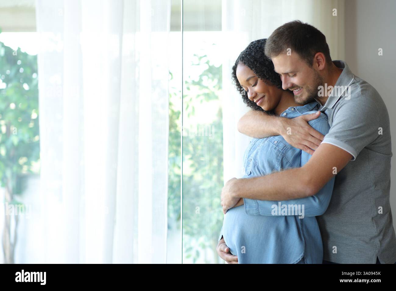 Happy interracial couple enjoying pregnancy together looking at belly at home Stock Photo