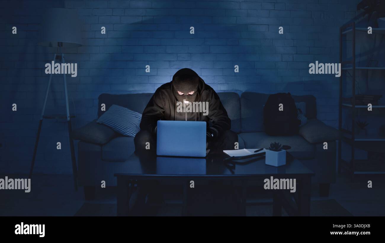 Masked hacker downloading private information using laptop Stock Photo