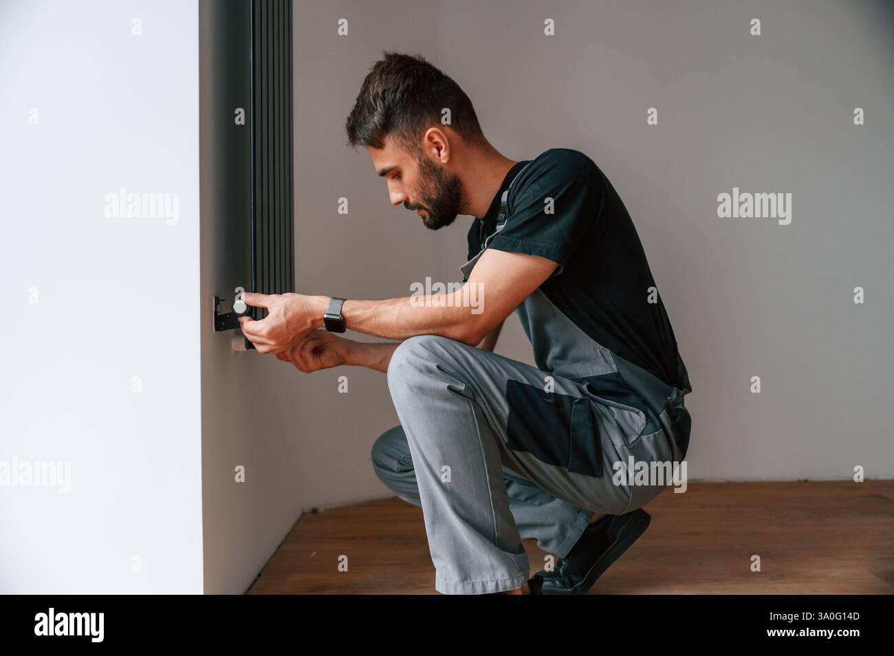 Plumber worker installing heating radiator in empty room of a newly built house. Stock Photo