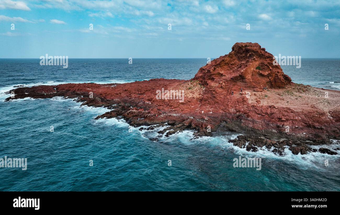 This captivating Socotra landscape features rugged red cliffs meeting tranquil blue waters against a cloudy sky. Stock Photo