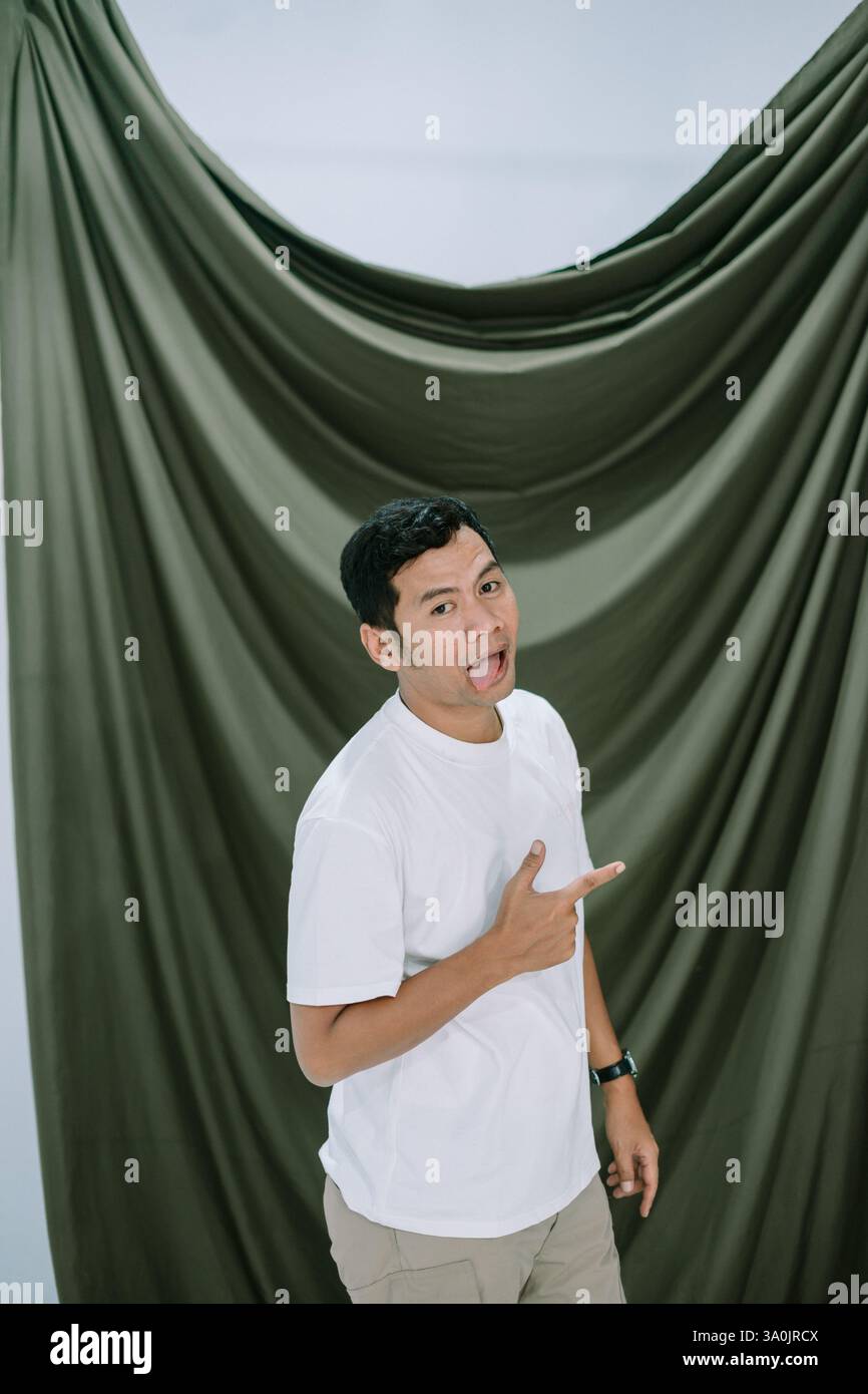 A young Asian man playfully sticks his tongue out and points to the side. Shot in studio with a draped green fabric backdrop. He's wearing a white t-s Stock Photo
