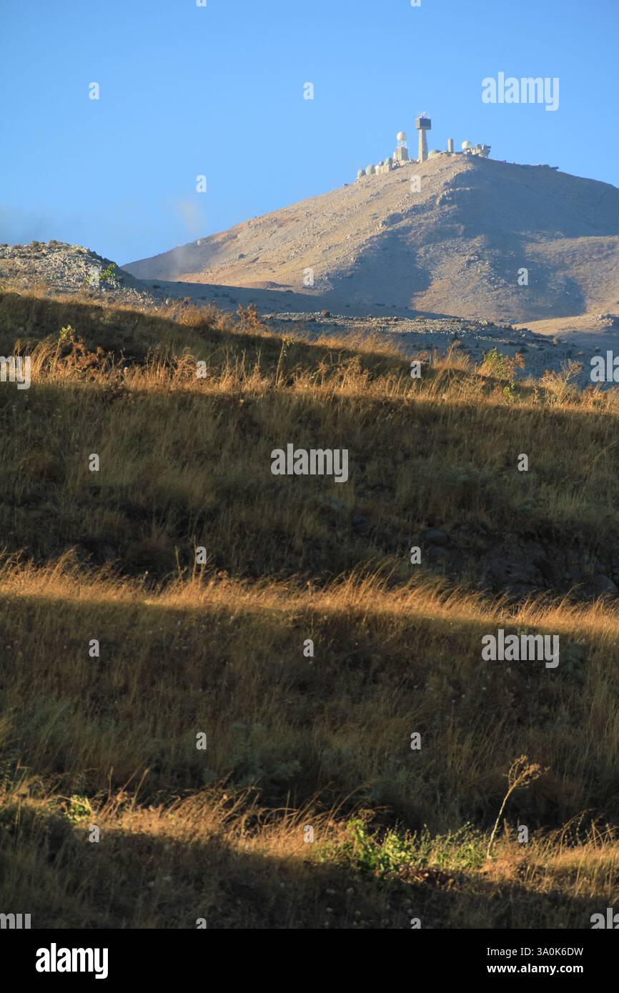 A scenic view of a mountain military radars  surrounded by dry grass and sparse trees in sunlight. The blue sky highlights the serenity and natural be Stock Photo