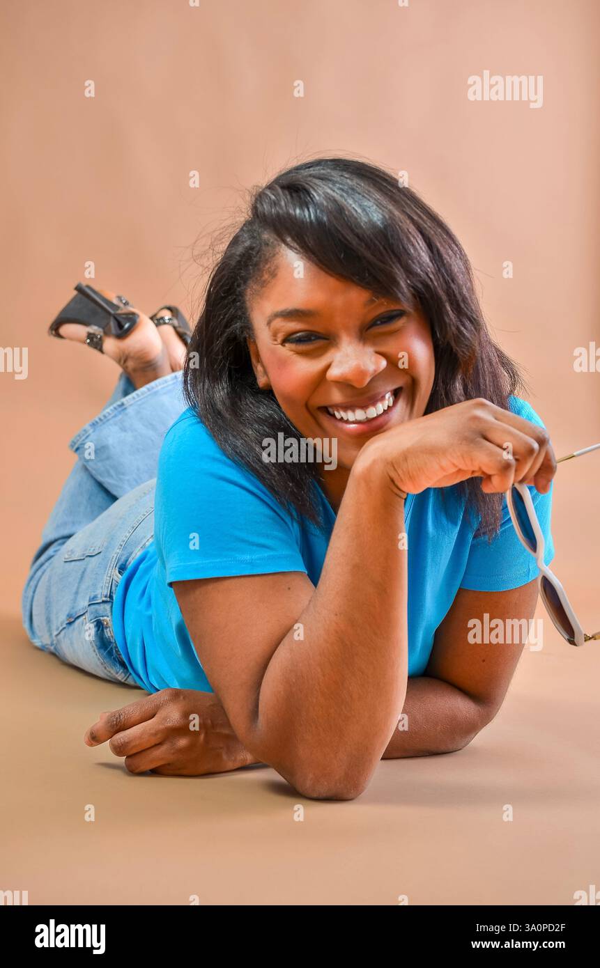 Studio portrait of a cheerful african american woman lying on the floor, holding sunglasses and smiling Stock Photo