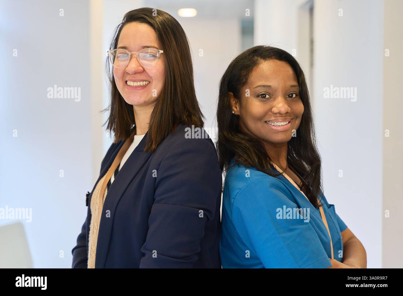 Portrait featuring a dentist and a manager standing back to back, both smiling confidently in a bright, modern dental clinic Stock Photo